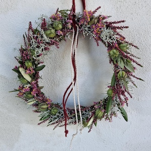 Peut inclure: Une couronne circulaire composée de bruyère violette séchée, de gousses de graines vertes et de lichen blanc argenté. L'arrangement est suspendu par une boucle en fil de laine rouge foncé et une cordelette blanche, créant un décor botanique texturé.