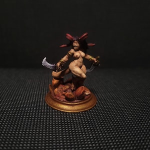 May include: A miniature figurine of a nude female warrior with dark hair and red accents, holding two curved blades. The figure is posed atop a detailed base with a gold-colored rim. The overall color palette is earthy tones.