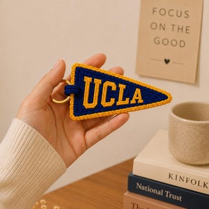 May include: A blue and gold UCLA pennant keychain. The keychain is triangular, with the letters "UCLA" in gold against a blue background. The pennant is trimmed with gold thread and attached to a gold keyring.