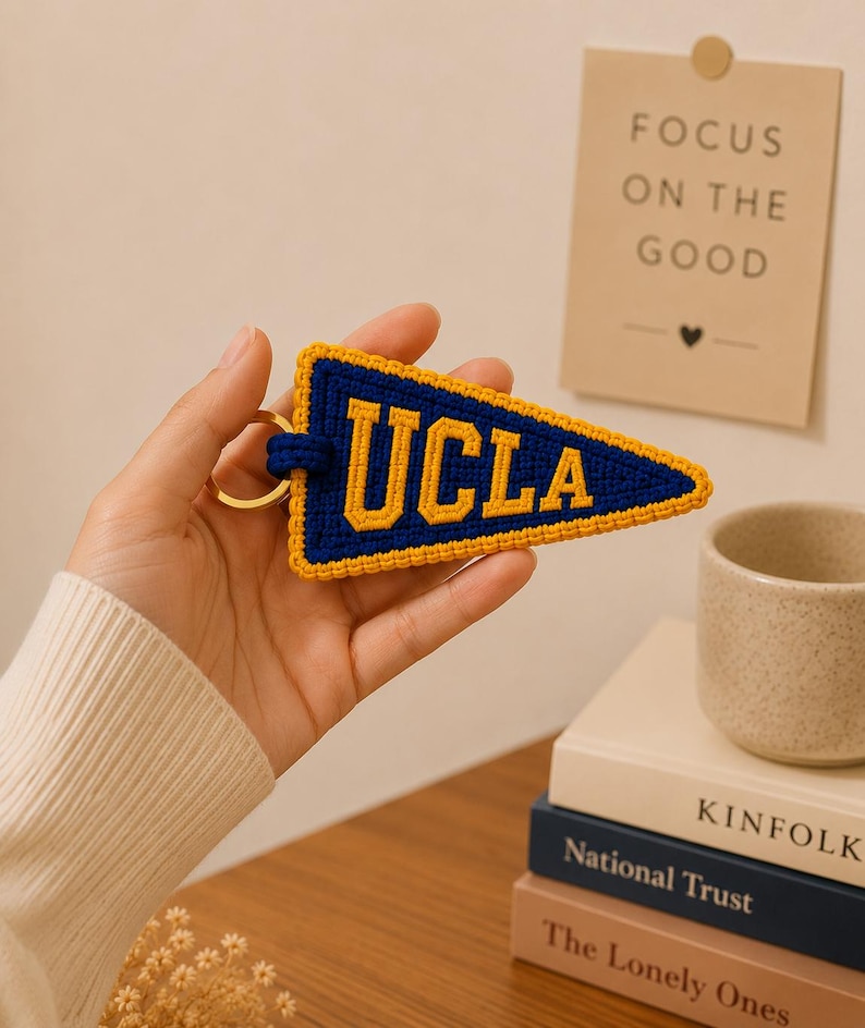 May include: A blue and gold UCLA pennant keychain. The keychain is triangular, with the letters "UCLA" in gold against a blue background. The pennant is trimmed with gold thread and attached to a gold keyring.