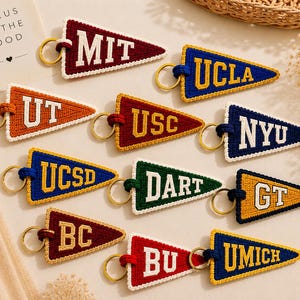 May include: A collection of colorful pennant-shaped keychains, each embroidered with a university's initials. The keychains feature a variety of colors, including red, blue, orange, and green, with gold-colored key rings. The text "FOCUS ON THE GOOD" is visible.