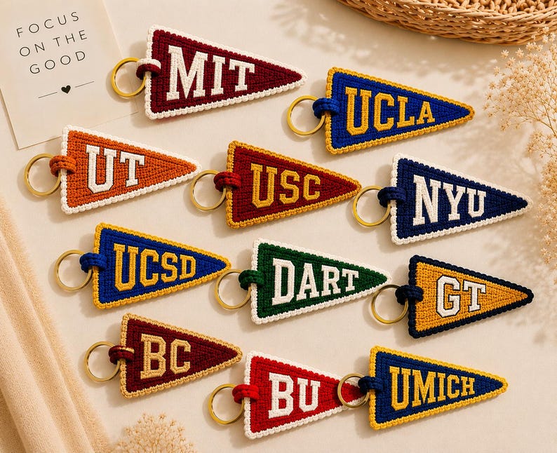 May include: A collection of colorful pennant-shaped keychains, each embroidered with a university's initials. The keychains feature a variety of colors, including red, blue, orange, and green, with gold-colored key rings. The text "FOCUS ON THE GOOD" is visible.
