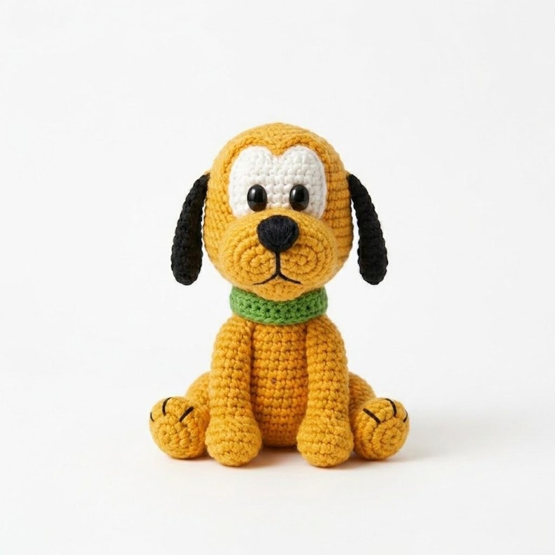 May include: A handmade crochet dog toy in a seated position. The toy features a yellow body, a white face, black ears, and a green collar. The dog has black button eyes and a black nose. The toy is made of yarn and sits on a white background.