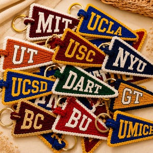 May include: A collection of colorful, handmade pennant keychains. Each keychain features a different university's initials, such as MIT, UCLA, and UT, in a contrasting color. The keychains are made of a textured material and have gold-colored key rings.