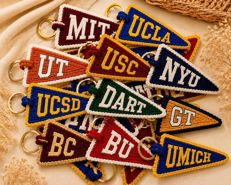 May include: A collection of colorful, handmade pennant keychains. Each keychain features a different university's initials, such as MIT, UCLA, and UT, in a contrasting color. The keychains are made of a textured material and have gold-colored key rings.