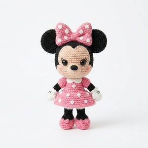 May include: A crocheted doll resembling a cartoon character. The doll has a beige face, black ears, and a pink bow with white polka dots. It wears a pink dress with white polka dots, black shoes, and white gloves. The doll is standing on a white surface.