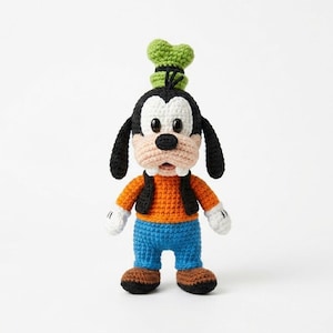 May include: A handmade crochet Goofy doll. The doll features a black and white head, black ears, and a green hat. It wears an orange shirt, blue pants, and brown shoes. The doll has white gloves and a black vest.