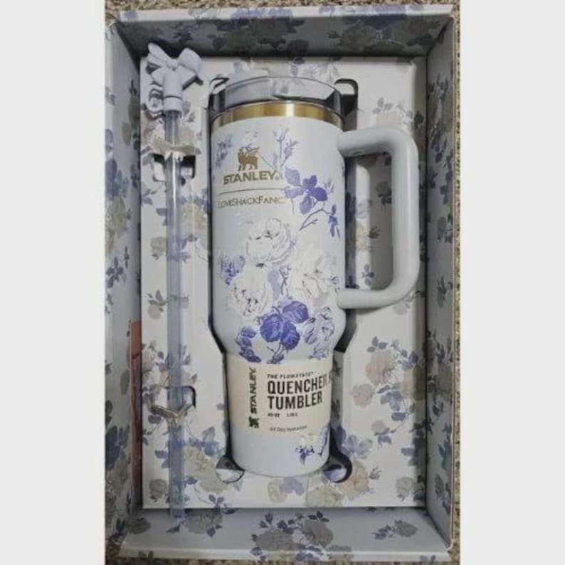 May include: A white Stanley Quencher tumbler with a blue floral pattern, a handle, and a matching straw in a box. The tumbler features the text Stanley, LoveShackFancy, and The Flowstate Quencher Tumbler 1.18 L. The box has a matching floral print.