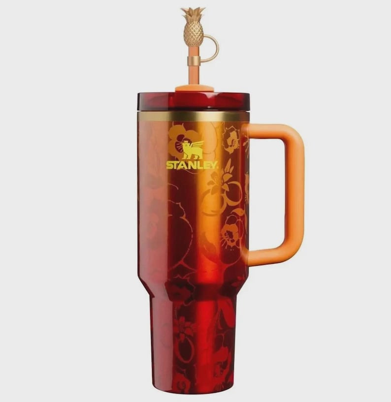 May include: A red and orange Stanley tumbler featuring a floral pattern, a gold-toned rim, and a matching orange handle. The lid includes a straw topped with a gold-colored pineapple charm. The Stanley logo is printed in yellow on the front.