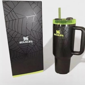 May include: A black Stanley tumbler with a lime green lid and straw, featuring a spiderweb pattern and the Stanley logo. Beside it stands a tall, rectangular black box decorated with a matching spiderweb design and the Stanley brand logo.