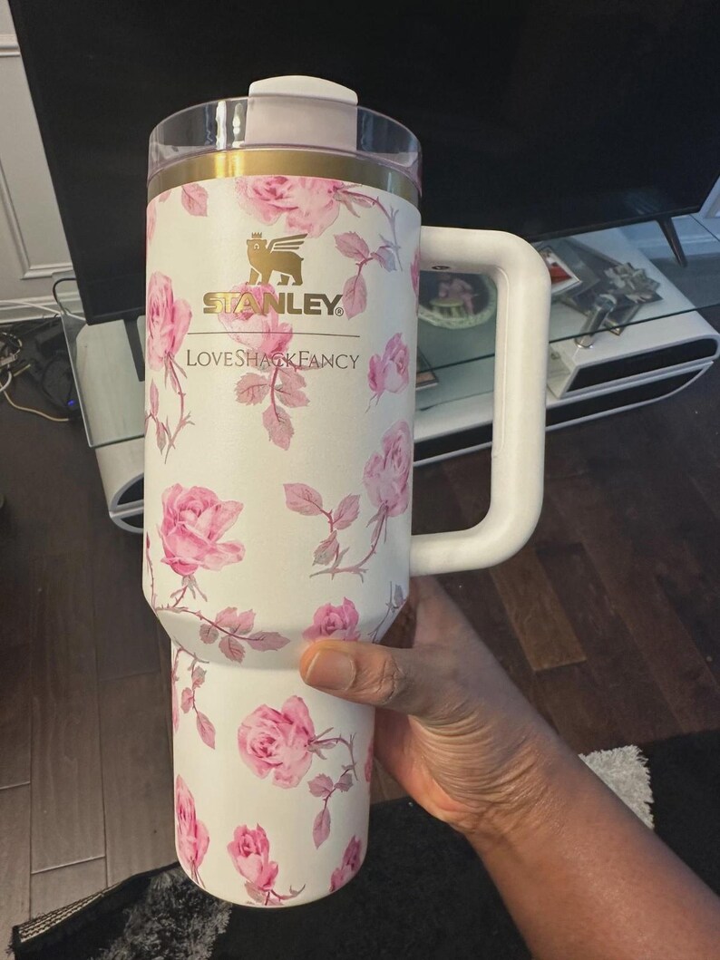 K&ouml;nnte beinhalten: Ein wei&szlig;er Isolierbecher mit einem rosa Rosenmuster, einem goldfarbenen Rand und einem transparenten Deckel. Auf der Vorderseite befinden sich das goldene Stanley-Logo und der Schriftzug LOVESHACKFANCY. Er hat einen gro&szlig;en Seitengriff.