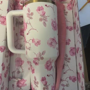 K&ouml;nnte beinhalten: Ein wei&szlig;er Isolierbecher mit einem rosa Rosenmuster, ausgestattet mit einem stabilen Seitengriff, einem transparenten Deckel mit goldfarbenem Rand und einem passenden durchsichtigen Strohhalm. Der Becher liegt in einer passend bedruckten Schachtel.
