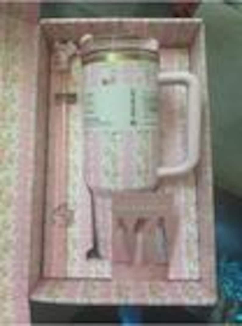 Puede incluir: Una taza rosa claro con estampado floral y tapa transparente, acompa&ntilde;ada de una cuchara mezcladora de mango largo a juego. El conjunto se presenta en una caja rectangular rosa con forro floral y una peque&ntilde;a tarjeta con tres figuras.