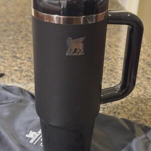 Puede incluir: Un vaso Stanley negro mate con una gran asa lateral y una tapa gris oscuro transl&uacute;cida. La taza presenta una banda met&aacute;lica plateada cerca de la parte superior y un logotipo Stanley plateado con un emblema de oso alado en el cuerpo cil&iacute;ndrico.