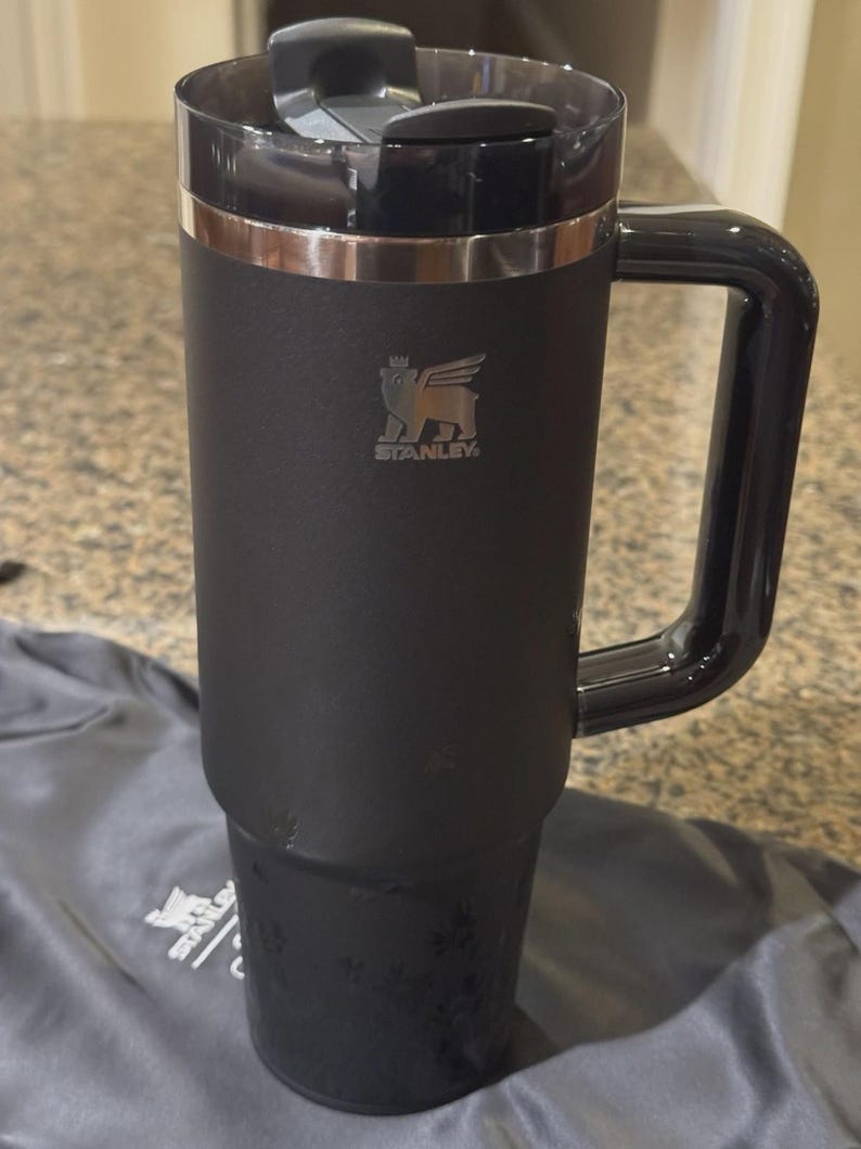 Puede incluir: Un vaso Stanley negro mate con una gran asa lateral y una tapa gris oscuro transl&uacute;cida. La taza presenta una banda met&aacute;lica plateada cerca de la parte superior y un logotipo Stanley plateado con un emblema de oso alado en el cuerpo cil&iacute;ndrico.