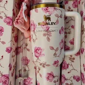 K&ouml;nnte beinhalten: Ein cremefarbener Stanley-Becher mit einem rosa Rosenmuster, der den Schriftzug Stanley und LoveShackFancy tr&auml;gt. Der Becher hat einen goldfarbenen Rand, einen wei&szlig;en Henkel, einen transparenten Deckel und einen passenden rosa Stoffbeutel.