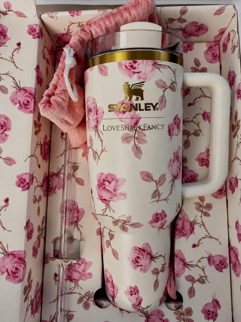 K&ouml;nnte beinhalten: Ein cremefarbener Stanley-Becher mit einem rosa Rosenmuster, der den Schriftzug Stanley und LoveShackFancy tr&auml;gt. Der Becher hat einen goldfarbenen Rand, einen wei&szlig;en Henkel, einen transparenten Deckel und einen passenden rosa Stoffbeutel.