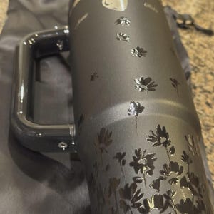 Puede incluir: Un vaso t&eacute;rmico negro mate con asa, que presenta un patr&oacute;n floral negro brillante de flores y p&eacute;talos dispersos. El dise&ntilde;o incluye una peque&ntilde;a forma de coraz&oacute;n cerca de la parte superior y un texto en cursiva que parece decir Ruby y James.