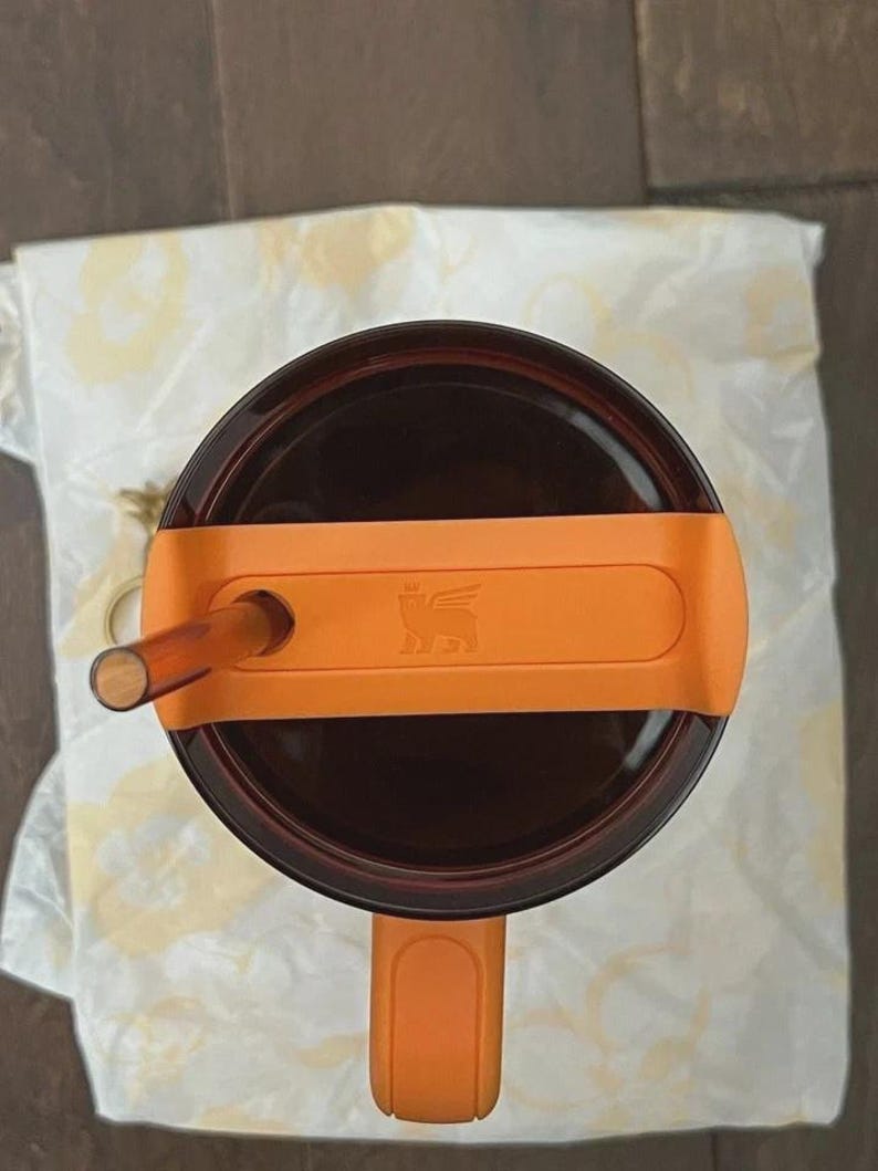 May include: A top-down view of a dark brown translucent tumbler with a bright orange lid and handle. The lid features a central orange bar with a small embossed winged bear logo and a circular opening holding a matching dark brown translucent straw.