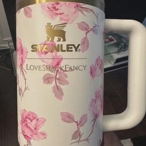 K&ouml;nnte beinhalten: Ein cremefarbener Isolierbecher mit einem rosa Rosenmuster. Auf der Vorderseite befindet sich ein goldenes Stanley-Logo mit einem gefl&uuml;gelten B&auml;rensymbol &uuml;ber dem Schriftzug LOVESHACKFANCY. Er verf&uuml;gt &uuml;ber einen transparenten Deckel, einen goldenen Rand und einen stabilen Seitengriff.