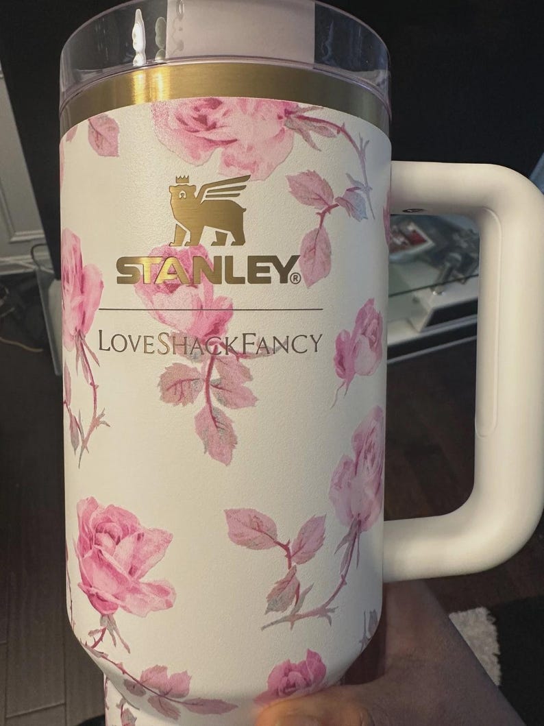 K&ouml;nnte beinhalten: Ein cremefarbener Isolierbecher mit einem rosa Rosenmuster. Auf der Vorderseite befindet sich ein goldenes Stanley-Logo mit einem gefl&uuml;gelten B&auml;rensymbol &uuml;ber dem Schriftzug LOVESHACKFANCY. Er verf&uuml;gt &uuml;ber einen transparenten Deckel, einen goldenen Rand und einen stabilen Seitengriff.