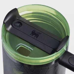 May include: A black travel mug featuring a translucent lime green lid with a black sliding closure mechanism. The lid displays a small embossed winged bear logo. The mug body is black with a subtle spider web pattern and a black handle.