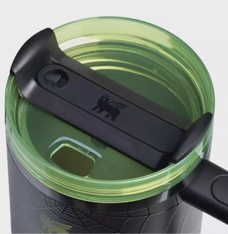 May include: A black travel mug featuring a translucent lime green lid with a black sliding closure mechanism. The lid displays a small embossed winged bear logo. The mug body is black with a subtle spider web pattern and a black handle.