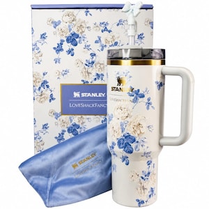 May include: A white Stanley LoveShackFancy tumbler with a blue and white floral pattern, a gold rim, a handle, and a bow-topped straw. It is accompanied by a matching floral box and a blue satin pouch featuring the Stanley LoveShackFancy logo.