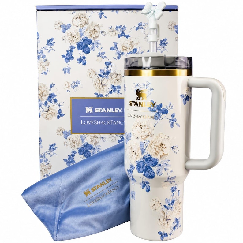May include: A white Stanley LoveShackFancy tumbler with a blue and white floral pattern, a gold rim, a handle, and a bow-topped straw. It is accompanied by a matching floral box and a blue satin pouch featuring the Stanley LoveShackFancy logo.
