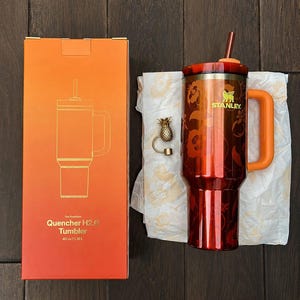 May include: A 40 oz Stanley Quencher H2.0 tumbler in a metallic orange finish with a floral pattern, a matching orange handle, and a straw. Beside it is a gold-toned pineapple charm and an orange box labeled The FlowState Quencher H2.0 Tumbler 40 oz | 1.18 L.