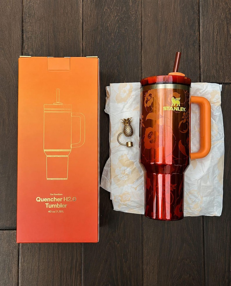May include: A 40 oz Stanley Quencher H2.0 tumbler in a metallic orange finish with a floral pattern, a matching orange handle, and a straw. Beside it is a gold-toned pineapple charm and an orange box labeled The FlowState Quencher H2.0 Tumbler 40 oz | 1.18 L.