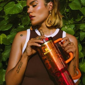 May include: A metallic orange Stanley tumbler featuring a floral pattern and a gold-toned pineapple-shaped topper on the lid. The insulated cup includes a sturdy orange handle and a wide base, showcasing the brand name Stanley in gold lettering.