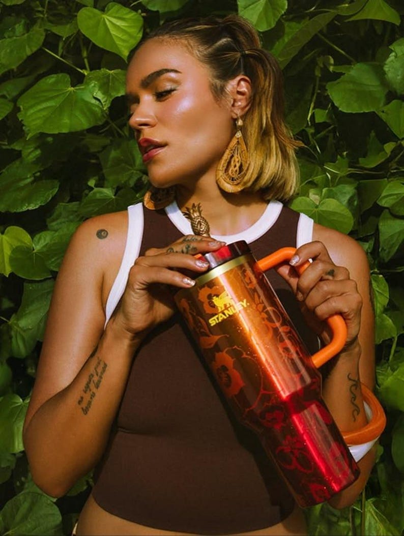 May include: A metallic orange Stanley tumbler featuring a floral pattern and a gold-toned pineapple-shaped topper on the lid. The insulated cup includes a sturdy orange handle and a wide base, showcasing the brand name Stanley in gold lettering.