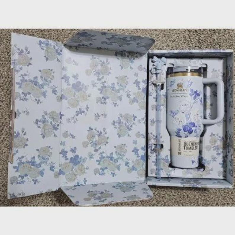 May include: A white Stanley Quencher tumbler with a blue floral pattern, a matching straw, and a handle, presented in a coordinating floral gift box. The tumbler features the Stanley logo and the text LoveShackFancy and Quencher Tumbler.