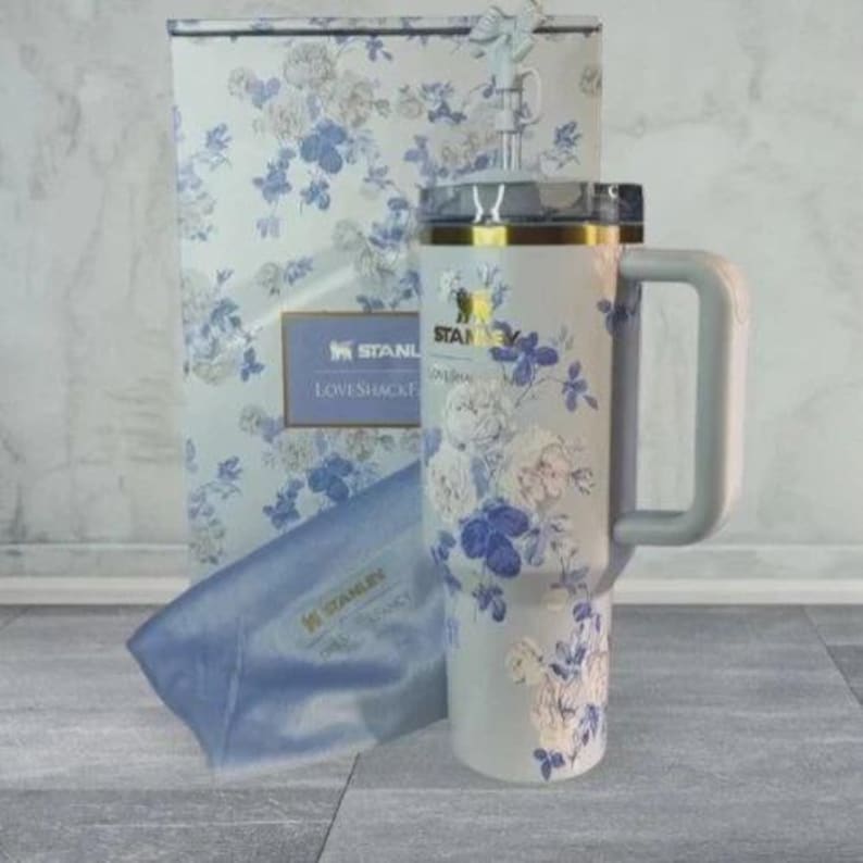 May include: A light grey Stanley tumbler with a blue and white floral pattern, a matching handle, and a clear lid with a decorative topper. The tumbler is displayed next to its original box, which features the Stanley and LoveShackFancy logos.