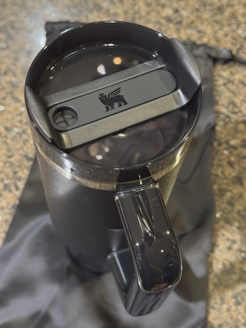 Puede incluir: Un vaso t&eacute;rmico negro mate con tapa y asa negras a juego. La tapa cuenta con un mecanismo de cierre deslizante con un peque&ntilde;o logotipo de un oso alado grabado en el centro. El vaso est&aacute; colocado en posici&oacute;n vertical, vista desde arriba.