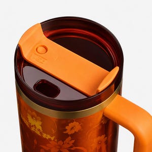 May include: A travel mug featuring a deep orange body with a floral pattern, a solid orange handle, and a dark translucent lid. The lid includes a sliding orange mechanism for the drinking opening, which is positioned on the top of the mug.