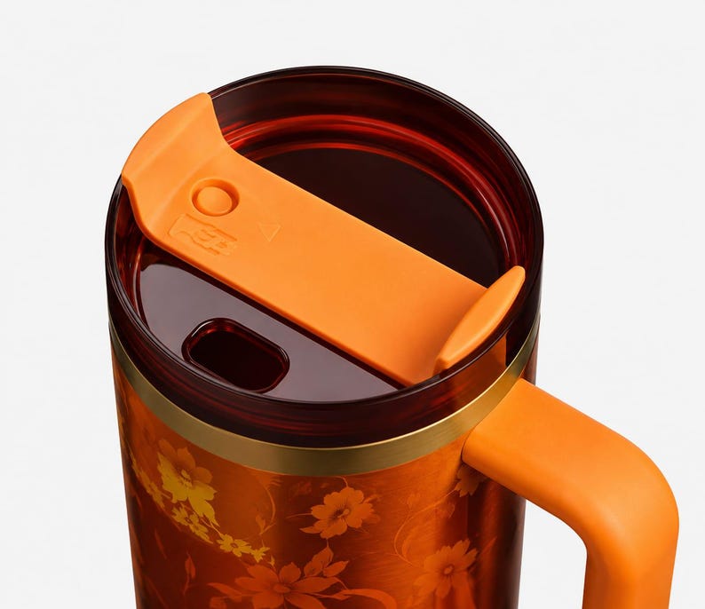 May include: A travel mug featuring a deep orange body with a floral pattern, a solid orange handle, and a dark translucent lid. The lid includes a sliding orange mechanism for the drinking opening, which is positioned on the top of the mug.
