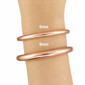 Puede incluir: Dos brazaletes de color cobre, de 6 mm y 8 mm de ancho, se muestran en una muñeca. Los brazaletes lisos y redondeados reflejan la luz, resaltando su brillo metálico. La imagen muestra la diferencia de tamaño entre los dos brazaletes.