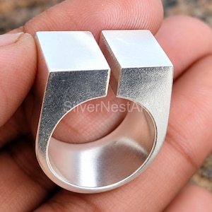 Könnte beinhalten: Ein silberfarbener Ring mit einem breiten, oben offenen Banddesign und zwei markanten, flachen quadratischen Blöcken an den Enden. Die Metalloberfläche hat ein glattes, mattes Finish und der Ring ist so ausgerichtet, dass die Lücke sichtbar ist.
