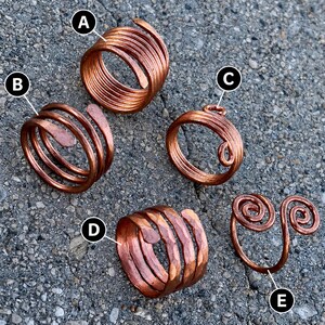 Puede incluir: Cinco anillos de cobre de varios diseños se muestran sobre una superficie gris. Los anillos incluyen diseños enrollados, en espiral y abiertos. El cobre tiene un tono marrón rojizo cálido y los anillos parecen hechos a mano.