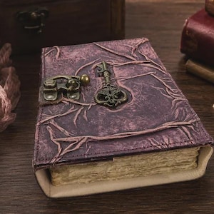 Peut inclure: Un journal rectangulaire avec une couverture violet foncé texturée présentant un motif en relief rappelant des branches. Le journal est fermé par un loquet en métal couleur laiton et orné d'une clé décorative en laiton sur le devant.