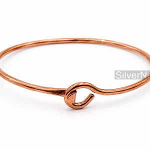Puede incluir: Una pulsera de cobre con una banda de alambre lisa y redondeada y un cierre de gancho. El metal tiene un acabado metálico cálido de color marrón rojizo con detalles de textura martillada cerca del cierre. El texto SilverNestArt es visible.