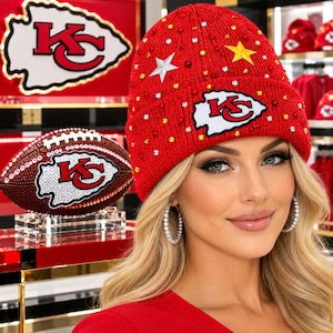 May include: A red knit beanie featuring the Kansas City Chiefs logo, embellished with scattered clear and yellow rhinestones and two star-shaped patches. A matching football with the same team logo and rhinestone detailing is displayed nearby.