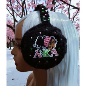 May include: Black faux fur earmuffs with the letters "AKA" embroidered in pink and green. The earmuffs have a decorative design with rhinestones and a silhouette of a woman's face. The headband is also black and decorated with rhinestones.