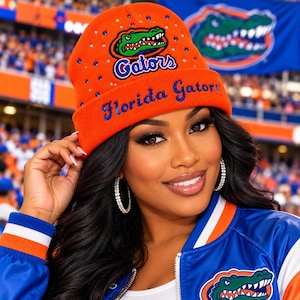 May include: An orange knit beanie featuring the Florida Gators logo, the text "Florida Gators" in blue glitter, and scattered blue and clear rhinestones. The beanie has a cuffed brim and is made of a soft, flexible knit material.