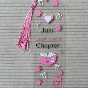 Puede incluir: Un marcapáginas transparente con una borla rosa, flores rosas y lazos blancos. El marcapáginas tiene el texto "Just one more Chapter" y presenta una ilustración de libro rosa con cerezas.