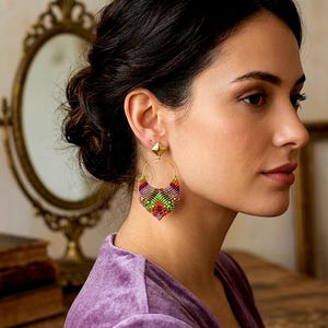 Puede incluir: Pendientes dorados con un perno cuadrado y un diseño tejido en forma de corazón en tonos morado, verde, naranja y amarillo. Los pendientes son un accesorio único, que añade un toque de color y textura.