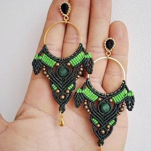 May include: Handmade macrame earrings featuring a dark green and lime green design with gold-colored accents. The earrings have a circular gold hoop and a teardrop-shaped charm. A black gem tops the earring.