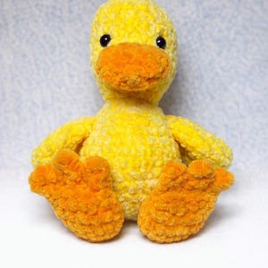 Puede incluir: Un pato de peluche amarillo hecho a mano con pico y patas naranjas. El pato tiene ojos negros y está sentado erguido. El peluche está hecho de un material suave y texturizado.